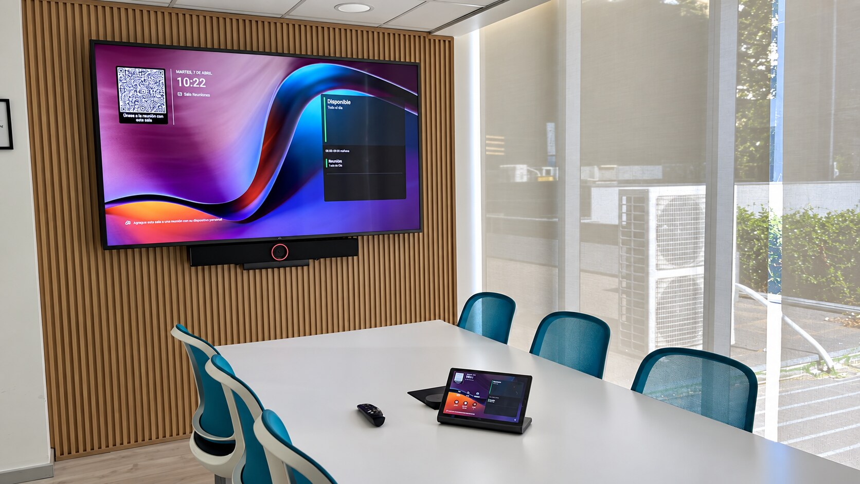 Sala de reuniones con pantalla mural, equipo de videoconferencia y panel táctil de mesa junto a un ventanal.