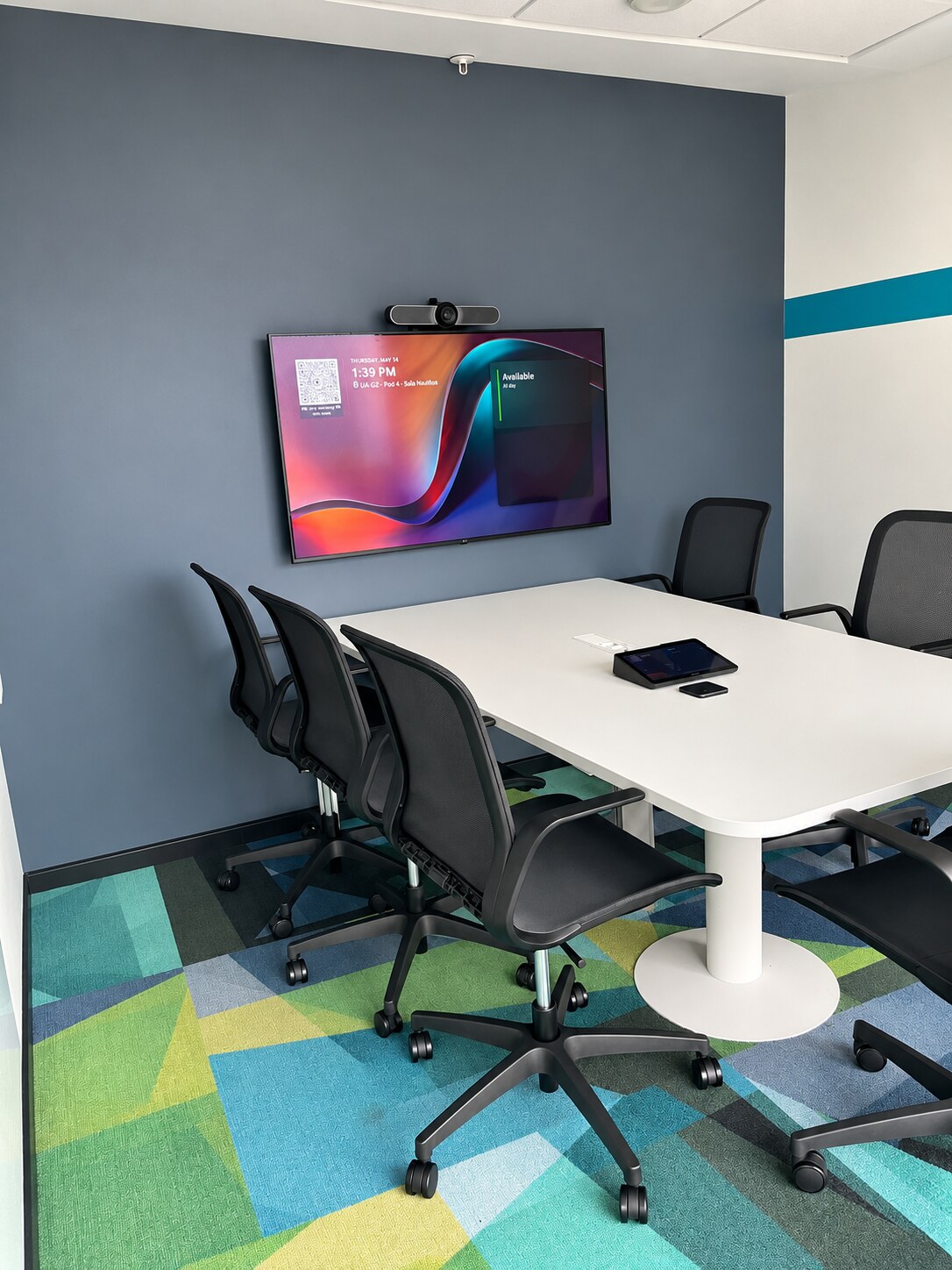 Sala de reuniones con pantalla mural, cámara de videoconferencia y panel táctil sobre la mesa.